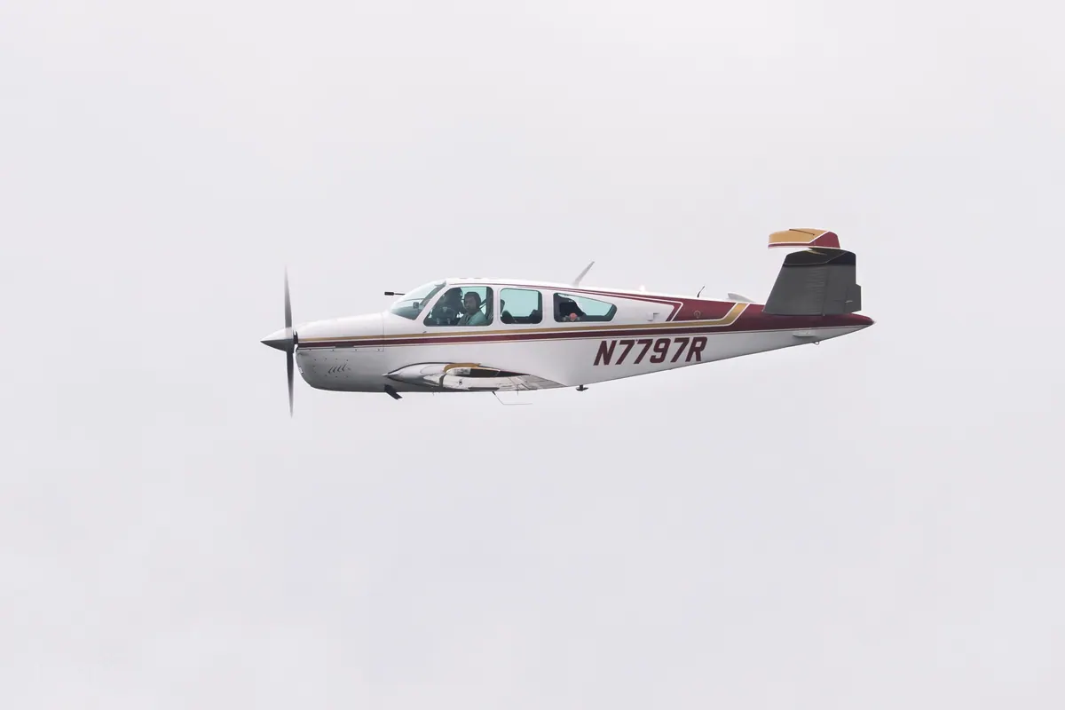 A white Beechcraft Bonanza with maroon and gold stripes, registration N7797R, flying level in profile against a flat white sky