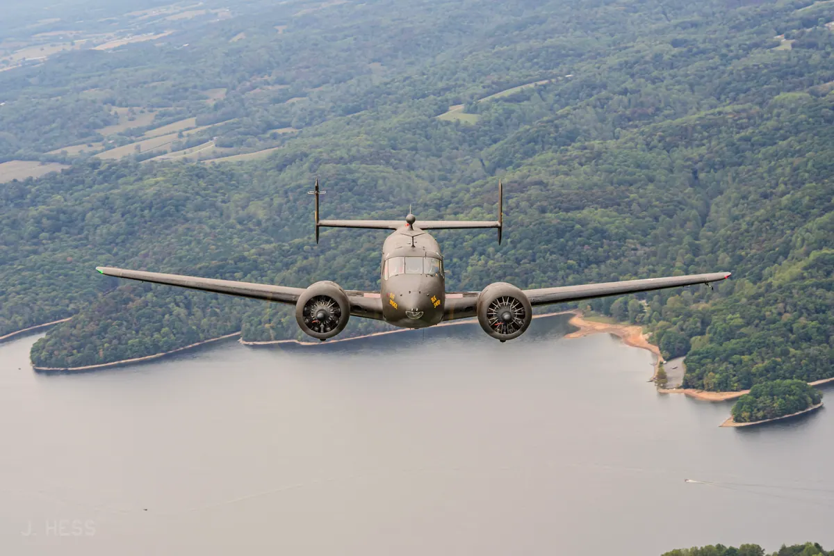 The olive-drab 'Bug Smasher' Beech 18 flies straight at the camera, wings level, above a wide lake ringed by forested hills